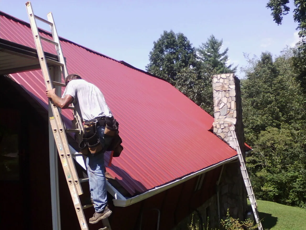 Roofer on a ladder beside a red metal roof and stone chimney — Soffit & Fascia Repair by Roofing Brothers in Mount Perry